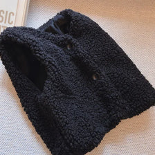 A black, chunky knit lamb wool vest with a V-neck and buttons is displayed on a light surface.