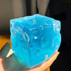 A hand holds a translucent blue ice cube shaped like a cube, used as a coconut oil scented squishy stress ball.