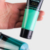 A hand holds a tube of emerald green acrylic color paint and an open tube of plastic paint with sealing film.