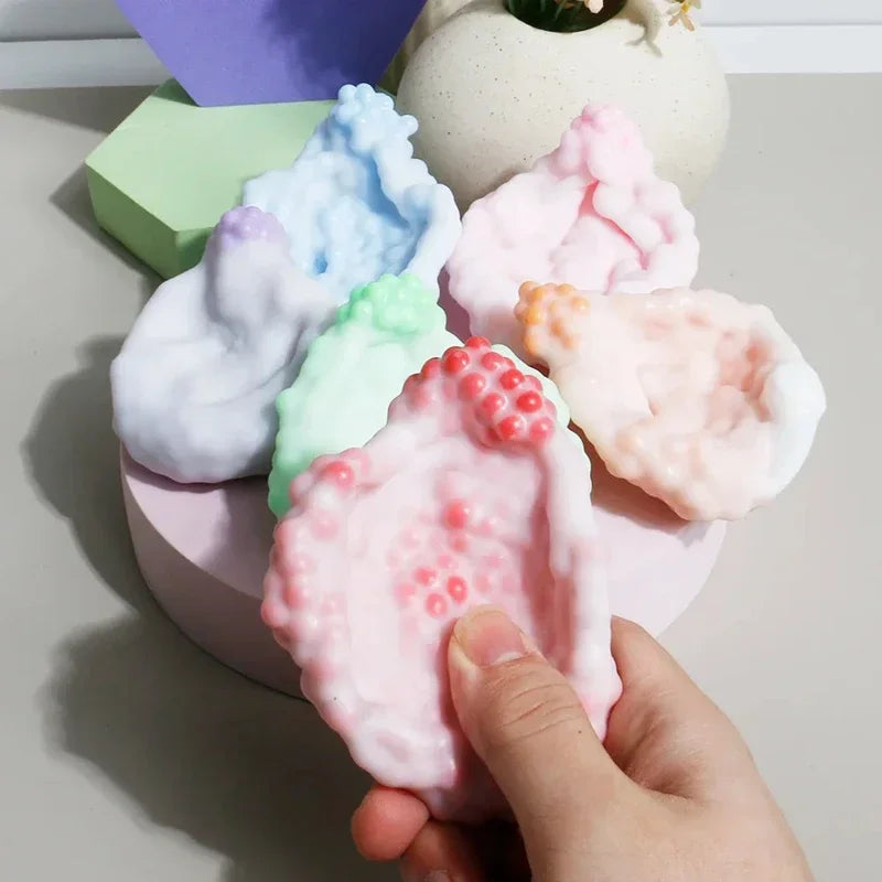A hand is squeezing a pink squishy stress relief ball with various pastel colored squishies in the background.