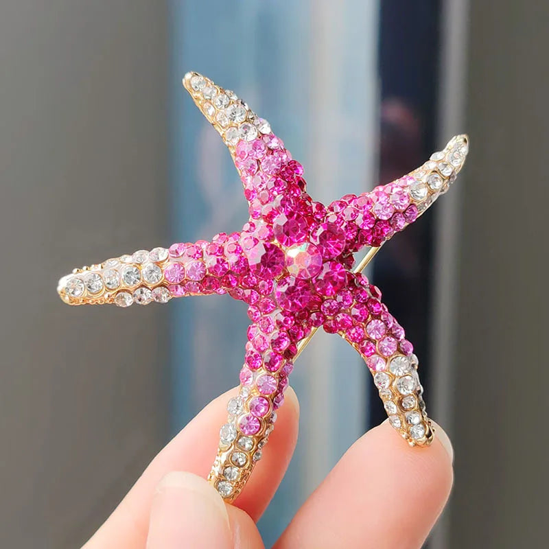 A pink and gold rhinestone turtle brooch pin is held by a person's fingers.
