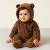 A baby wearing a brown fleece hooded romper with a zipper, sitting on a light surface.