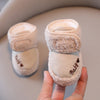 A hand holds a pair of light pink plush baby snow boots with a soft texture and anti-slip sole.