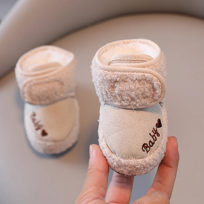 A hand holds a pair of light pink plush baby snow boots with a soft texture and anti-slip sole.