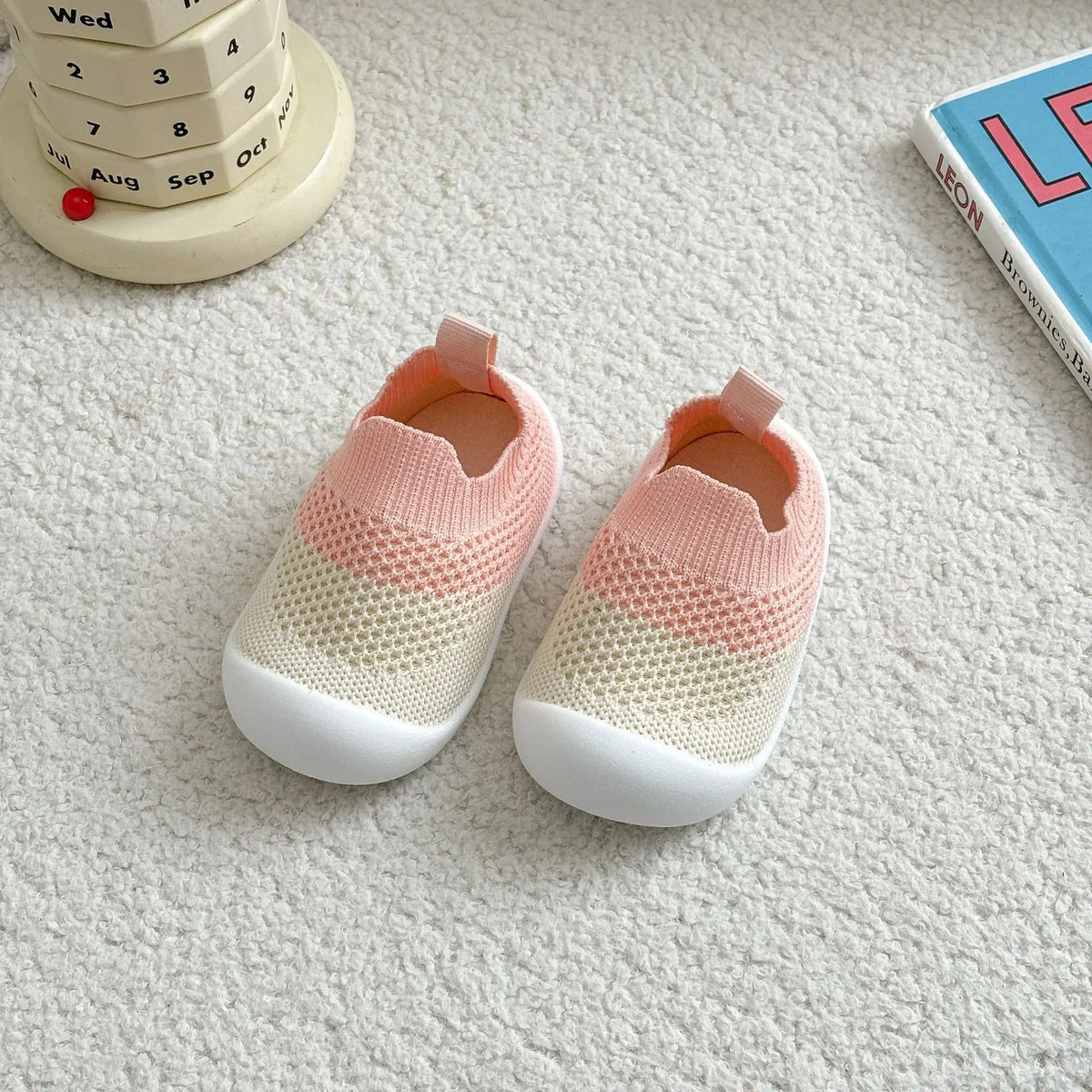 A pair of pink and cream breathable anti-slip baby first walker shoes resting on a white surface next to a wooden stacking to