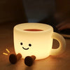 A white, coffee cup-shaped rechargeable night light with a smiling face and small arms rests on a wooden surface.