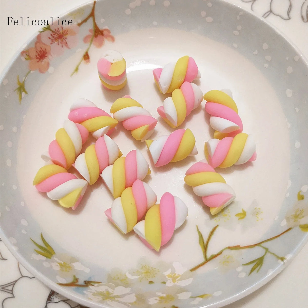 A collection of ten polymer clay marshmallow charms in pink and yellow stripes arranged on a white plate.