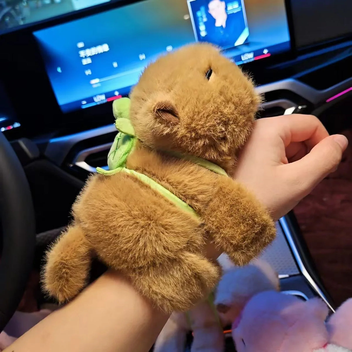 A hand is holding a brown capybara plush toy with a green strap, resting on a car dashboard.