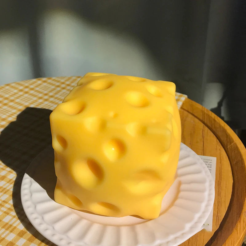 A yellow, squishy silicone fidget toy shaped like a cube is displayed on a white plate on a wooden surface.