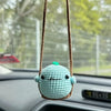 A light blue crocheted animal with a green accent is hanging from a brown string on a car interior.