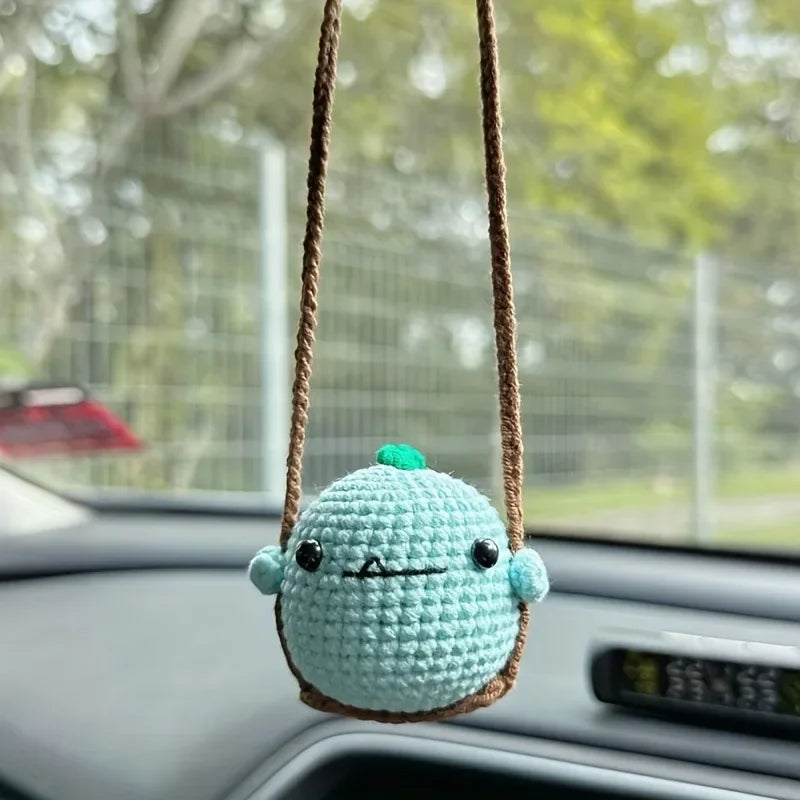 A light blue crocheted animal with a green accent is hanging from a brown string on a car interior.
