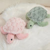 A pink sea turtle plush toy resting on a white fluffy surface next to a green sea turtle plush.