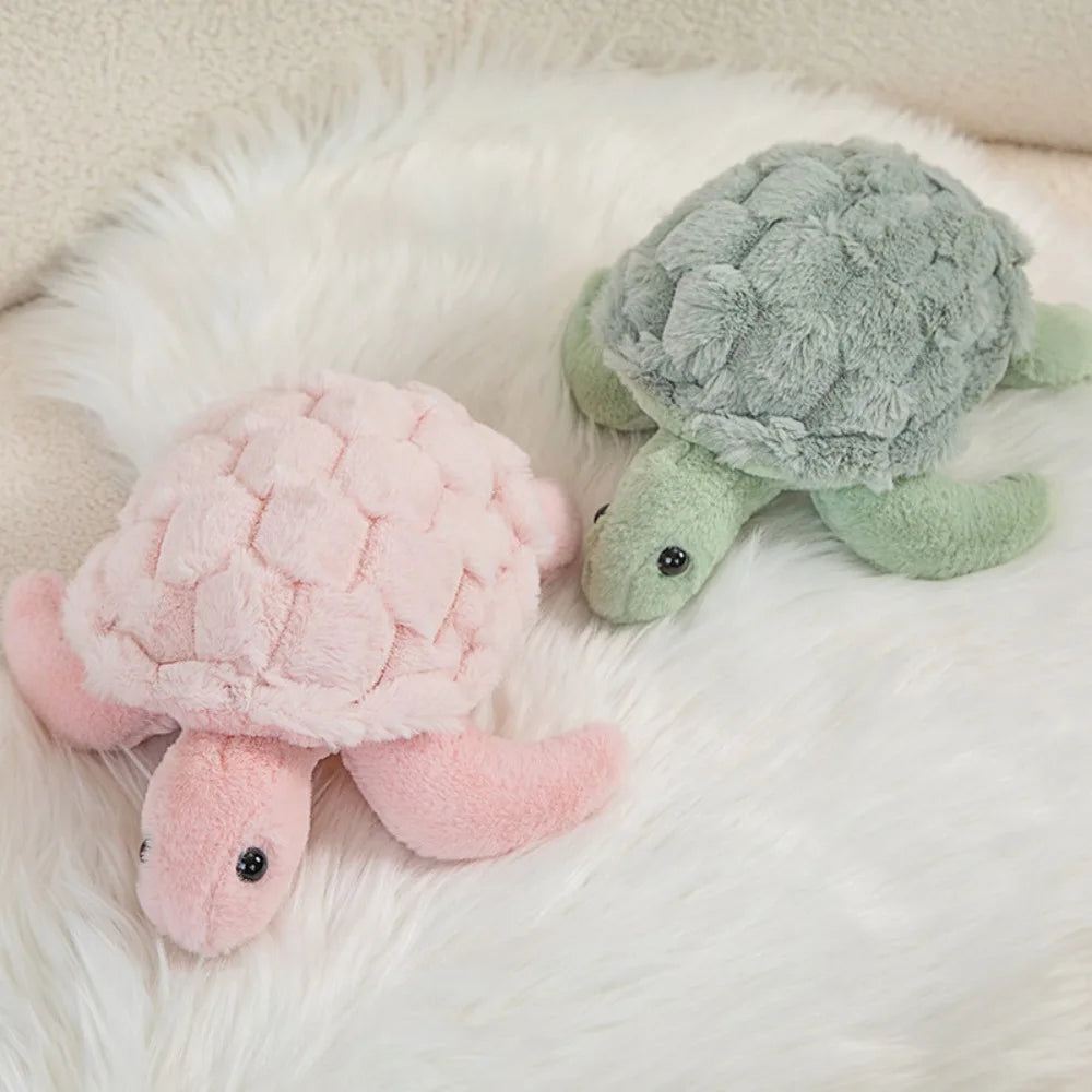 A pink sea turtle plush toy resting on a white fluffy surface next to a green sea turtle plush.