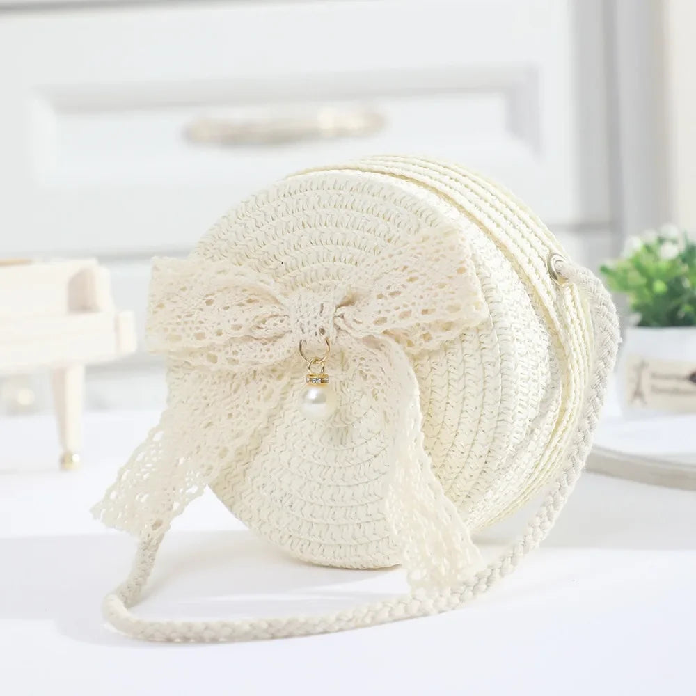 A cream-colored handwoven straw crossbody bag featuring a delicate white lace bow detail.
