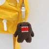 A brown, monster-shaped plush keychain charm with a red tooth detail hanging from a yellow fabric pouch.
