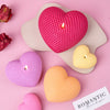 A set of three silicone heart candle molds in pink, yellow, and orange resting on a light pink surface.