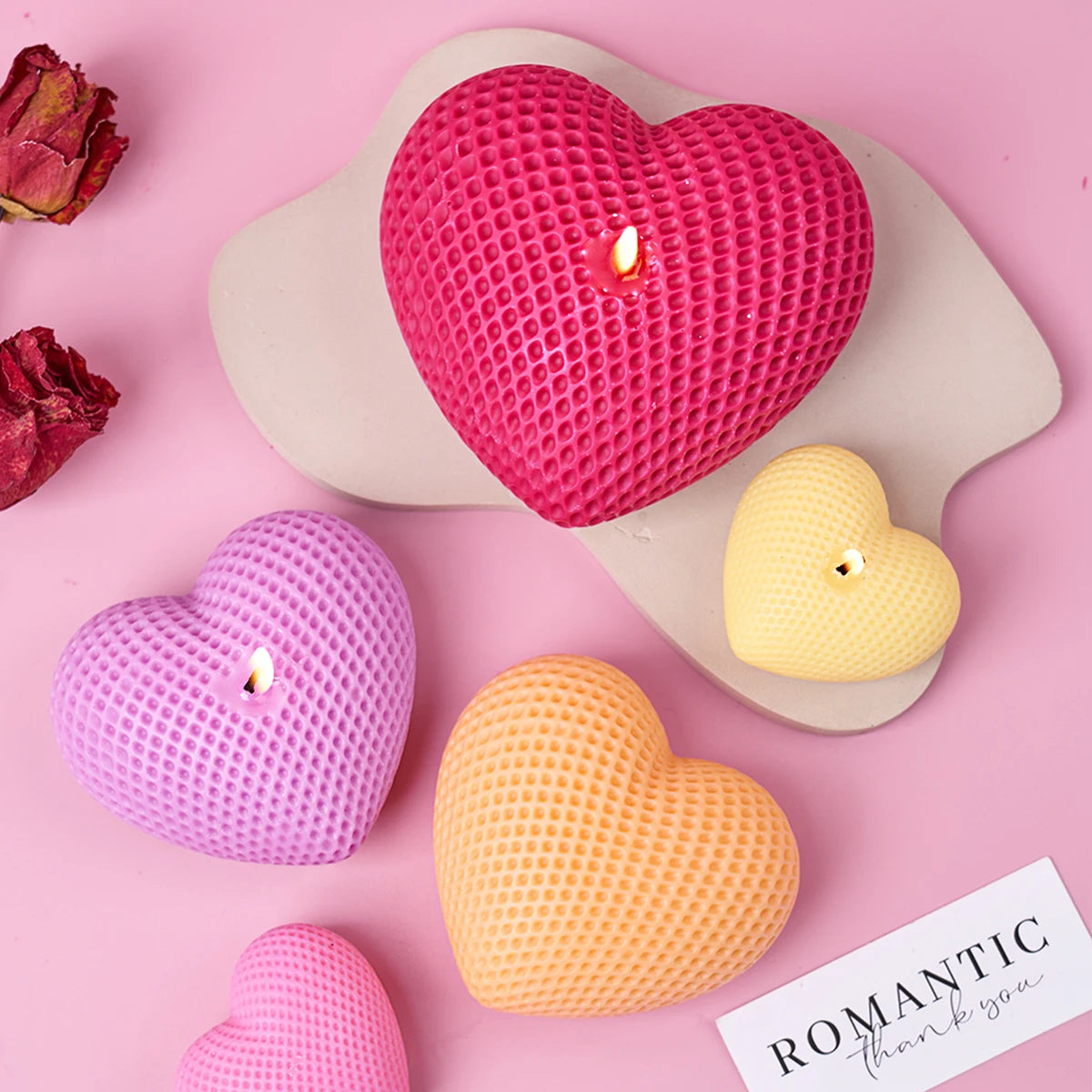 A set of three silicone heart candle molds in pink, yellow, and orange resting on a light pink surface.