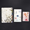 Three epoxy resin notebook covers featuring mechanical gears, starfish, and floral elements on white and pink backgrounds.