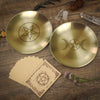 A brass pentagram altar plate rests on wood next to ritual parchment and pine cones.