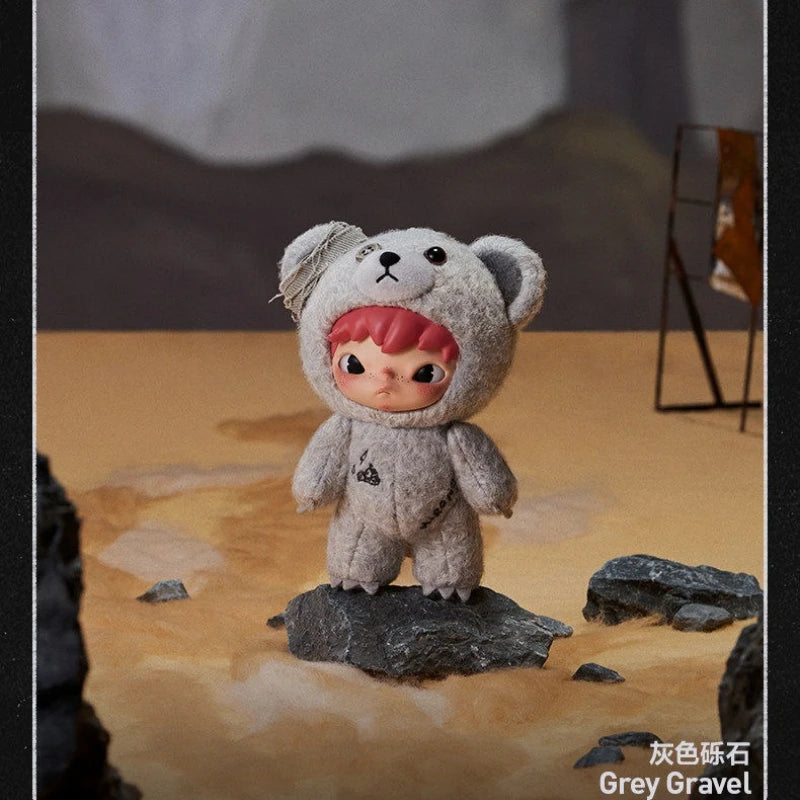 A small grey bear plush doll standing on a rock in a desert-like setting.