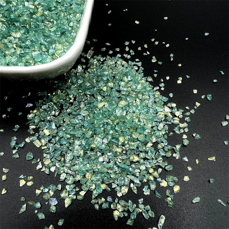Crushed green glass crystals scattered on a dark surface with a small white bowl nearby.