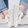 A pair of white kids barefoot sneakers with double hook-and-loop straps worn on a person's feet.