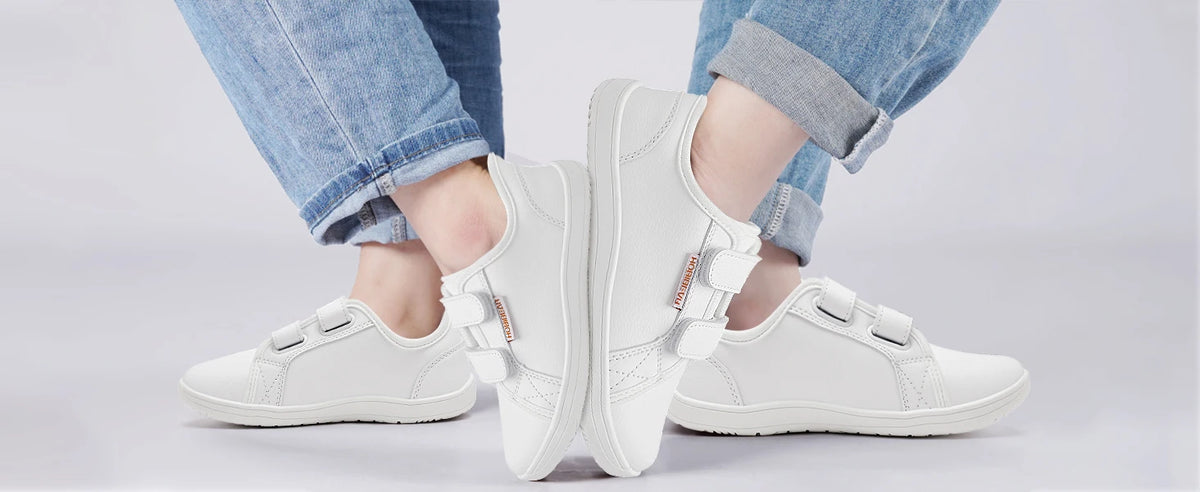 A pair of white kids barefoot sneakers with double hook-and-loop straps worn on a person's feet.
