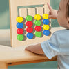 A baby is interacting with a wooden color and shape matching game board featuring colorful circular blocks.