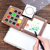 A portable wooden watercolor paint set with various colored paints and a blank canvas is shown on a wooden surface.