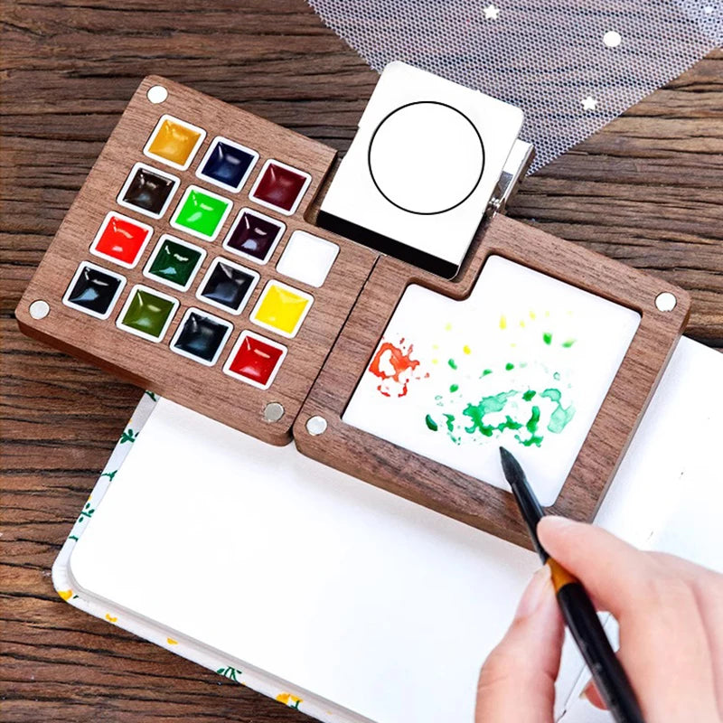 A portable wooden watercolor paint set with various colored paints and a blank canvas is shown on a wooden surface.