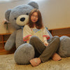 A young woman is sitting on the floor with a giant gray plush teddy bear and soft, light-colored pants.