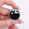 A hand holds a black enamel pin shaped like a cat's face with white markings.