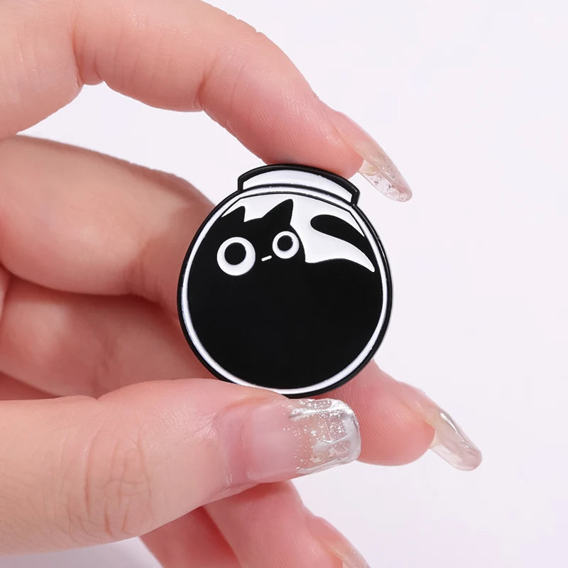 A hand holds a black enamel pin shaped like a cat's face with white markings.