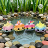 A set of eight colorful flexible salamander figurines resting in clear water surrounded by smooth stones and green aquatic pl