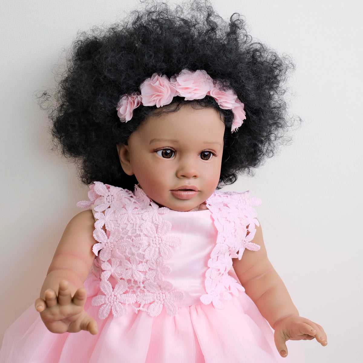A lifelike reborn toddler doll with hand-rooted black hair and a pink lace dress.