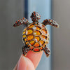 A detailed rhinestone turtle brooch pin with an amber shell and textured head is held by a hand.