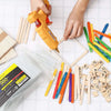 A hand is holding a yellow hot glue gun, with various colored and natural wood craft sticks scattered on a white surface.
