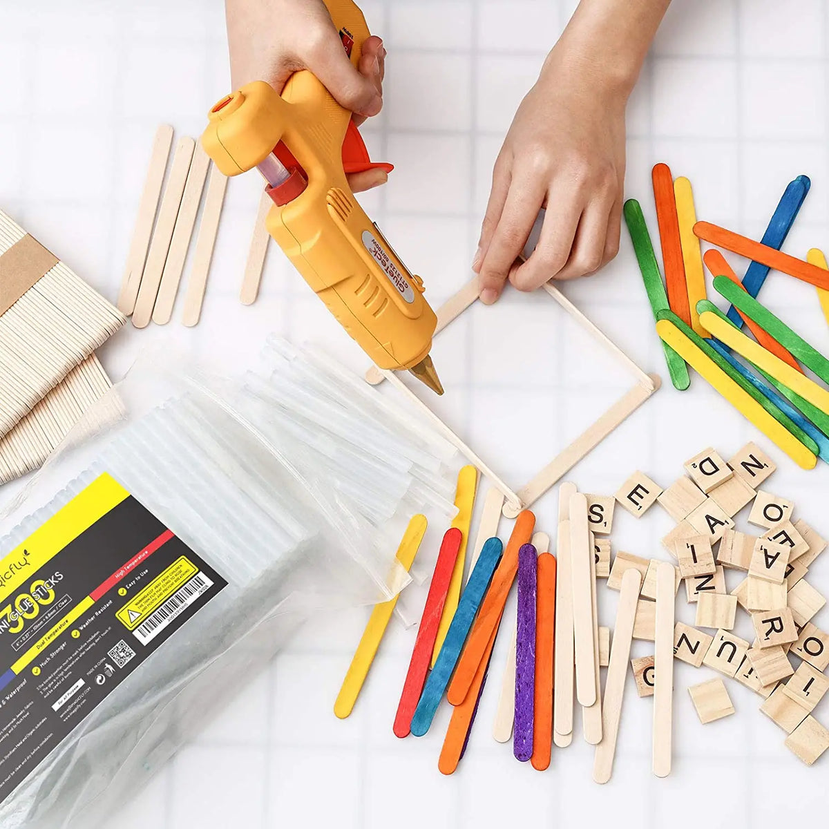 A hand is holding a yellow hot glue gun, with various colored and natural wood craft sticks scattered on a white surface.