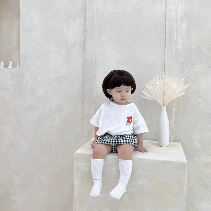 A young girl wearing a white t-shirt with a small floral print and plaid shorts, paired with white socks, sitting on a light 