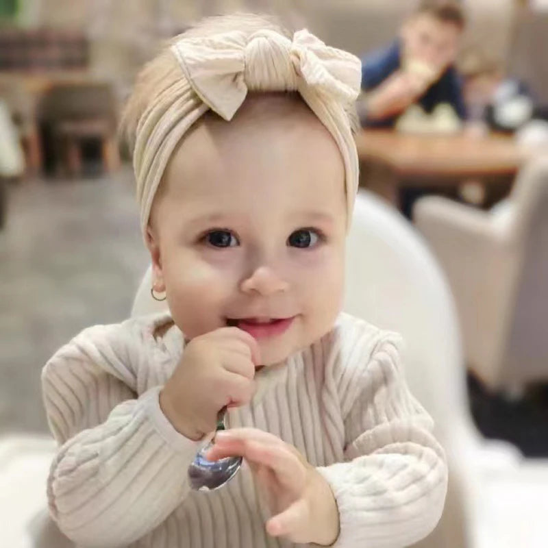 A baby wearing a beige soft stretch headband with a bow, smiling and touching their face.