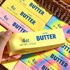 A hand holds a yellow butter stick fidget toy next to other salted butter sticks in a woven basket.