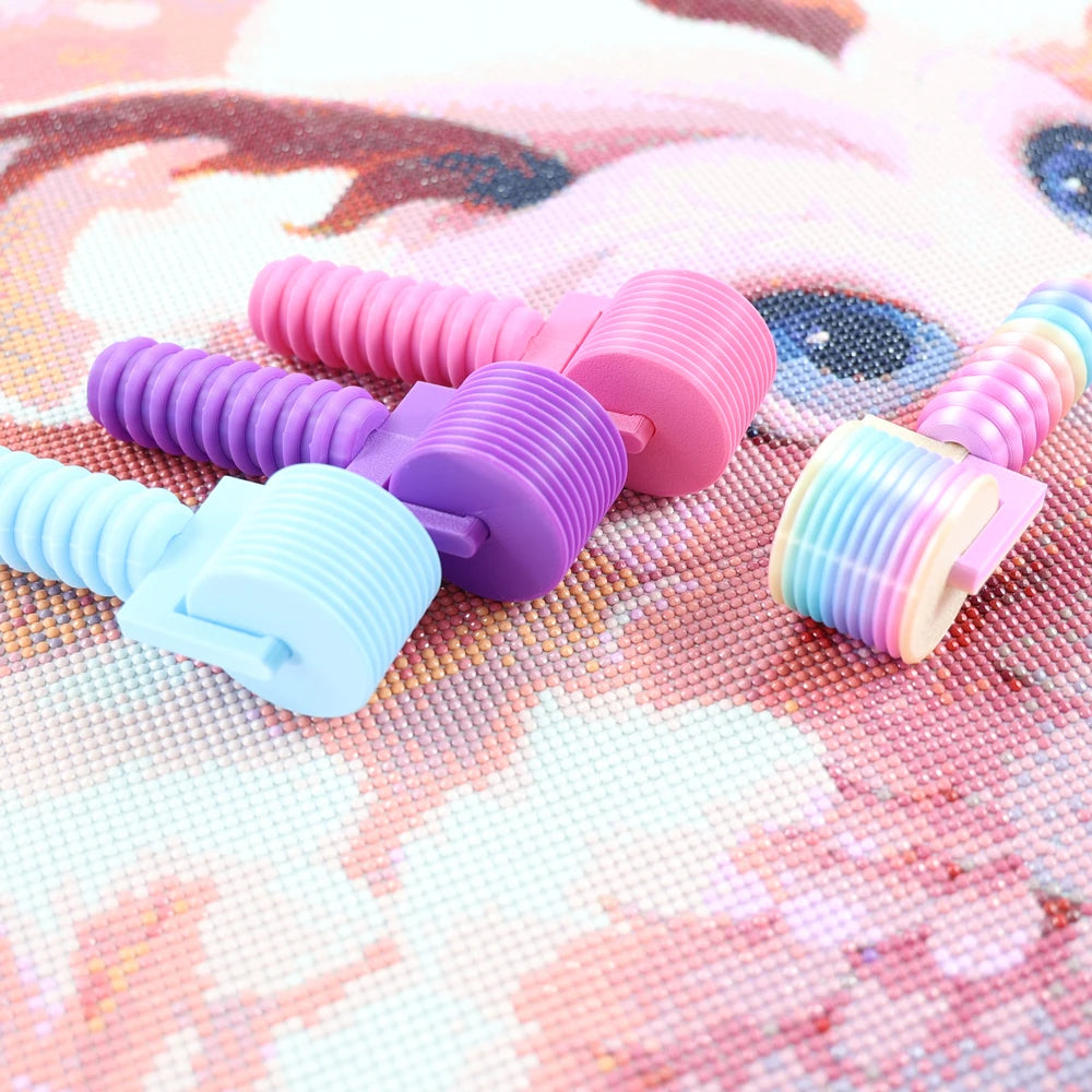 A collection of colorful, detachable diamond painting rollers in pink, purple, and blue are displayed on a pink patterned sur