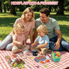 A family is enjoying a Kids Playfood and Picnic Set outdoors with fruits, vegetables, and cookware on a checkered blanket.