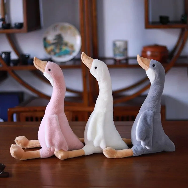 Three long neck goose plush dolls in pink, white, and gray sitting on a wooden table.