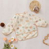 A light beige baby bib with long sleeves featuring a pattern of soft peach and green floral shapes, placed on a white linen b