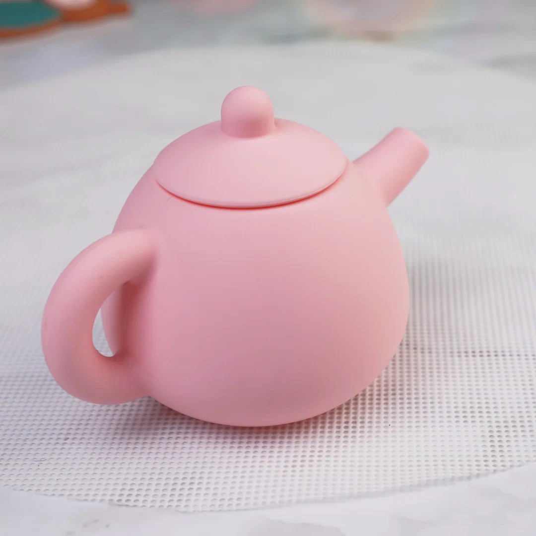 A pink silicone pretend play tea set teapot with a lid and spout resting on a white surface.