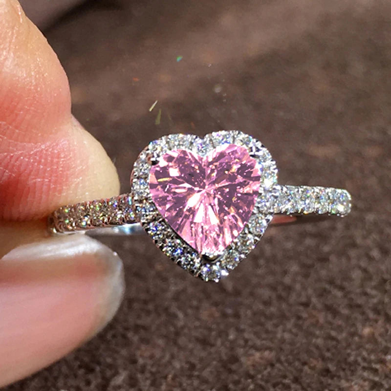 A close-up of a heart-shaped pink gemstone ring set in a halo of small diamonds being held by a person's finger.