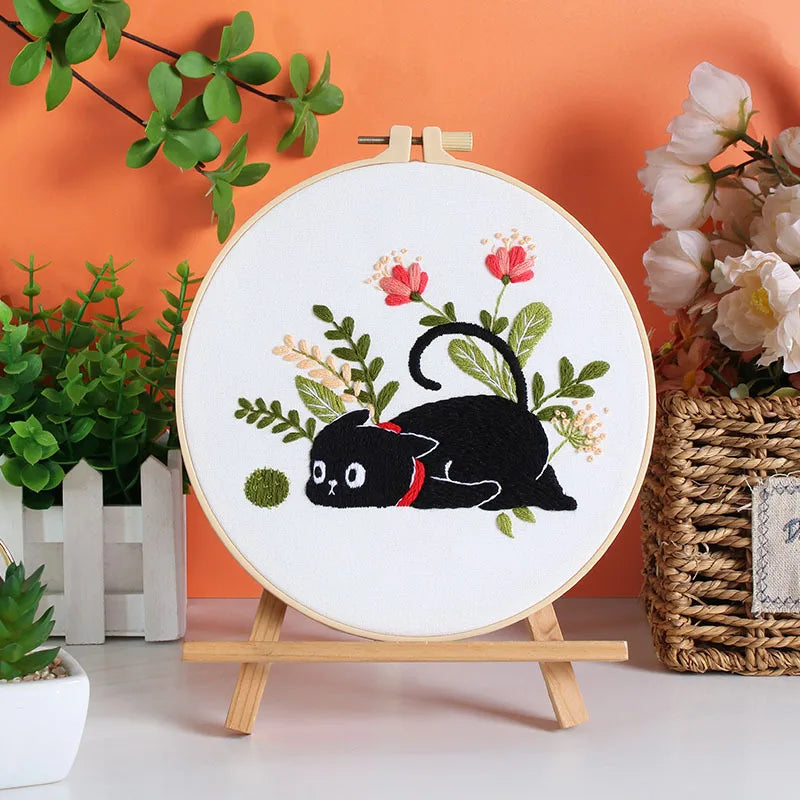 A white embroidery kit featuring a black cat design on a round canvas, displayed against an orange wall.