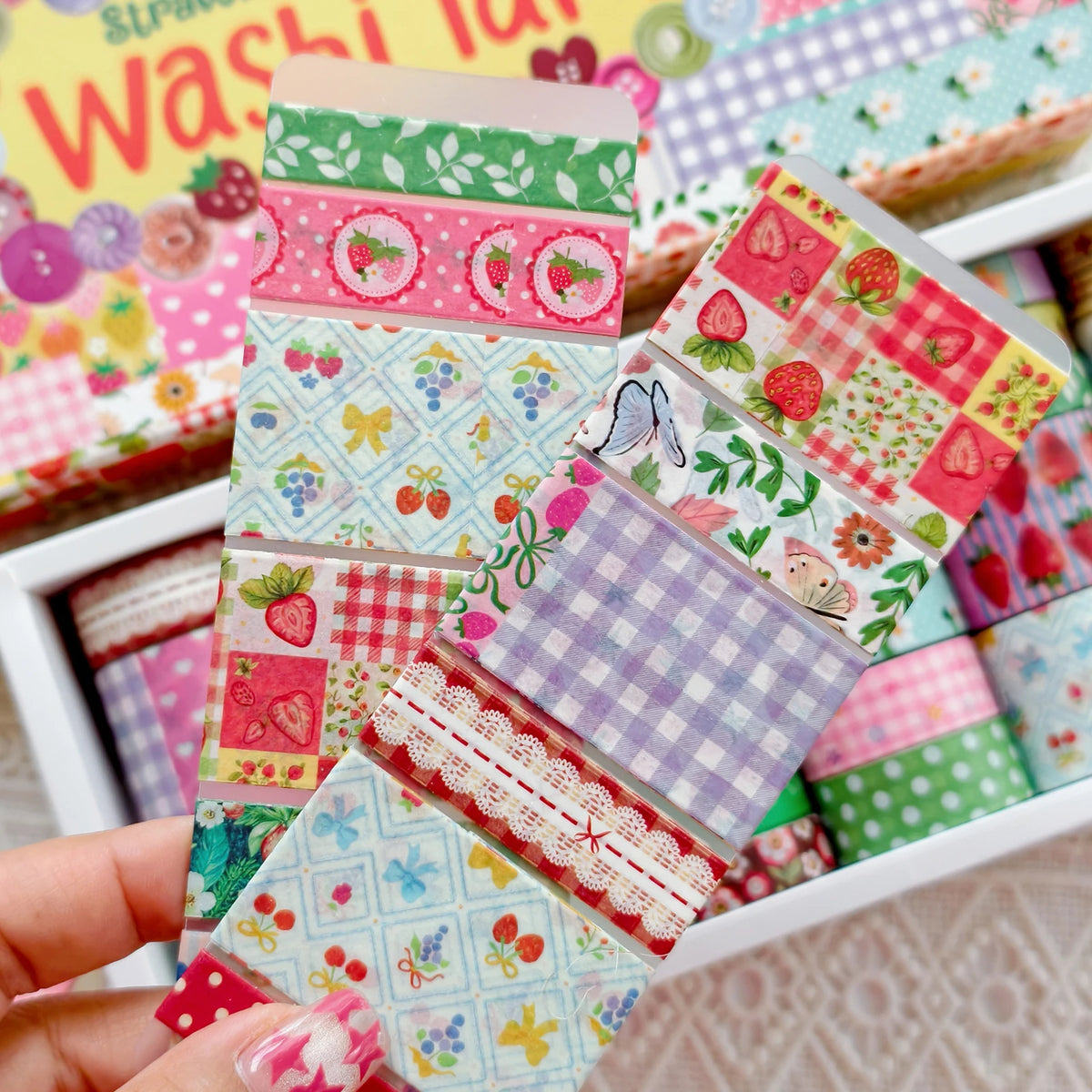 A hand holds a collection of colorful strawberry patterned washi tape rolls in a white box.