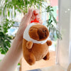 A hand holds a brown capybara plush toy with a white collar and a red charm against a blurred green background.